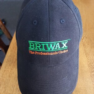May include: A black baseball cap featuring the embroidered green text BRIWAX with the orange text The Professionals Choice underneath. The cap has a structured crown with six panels, a button on top, and a curved brim, displaying a clean design.