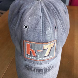 May include: A faded grey baseball cap featuring a rectangular patch with the orange and white text HT and the words hot tuna below it. The brim displays the word gumpz in a dark grey font. The cap has a metal button on top and metal eyelets.