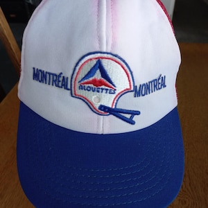 May include: A white and blue baseball cap featuring an embroidered logo of a football helmet with the text "MONTRÉAL ALOUETTES" and "MONTRÉAL" on the front. The cap has a solid blue brim, a white front panel, and a red mesh back with a red button on top.