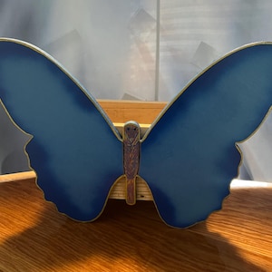 May include: A large, blue butterfly decoration with gold trim. The butterfly's wings are spread wide, and the body is brown. The decoration is made of wood and sits on a wooden surface.