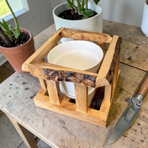May include: A wooden pet bowl holder with a natural bark edge. The holder is square with vertical supports and holds a white ceramic bowl. The bowl is filled with water. The holder sits on a wooden table.