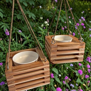 Könnte beinhalten: Zwei hängende Holzpflanzgefäße mit hellfarbigen Keramikschalen im Inneren. Die Pflanzgefäße bestehen aus horizontalen Holzlatten und sind an Seilen aufgehängt. Der Hintergrund zeigt grünes Laub und lila Blumen.