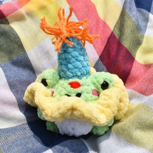 May include: A handmade crochet frog toy with a blue cone-shaped hat topped with orange yarn. The frog is green with yellow and white accents, and black eyes. The toy is sitting on a plaid blanket.