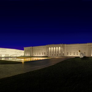 May include: Exterior shot of a modern art museum at dusk. The building is illuminated with warm lights, reflecting in a pool of water. The sky transitions from deep blue to purple.