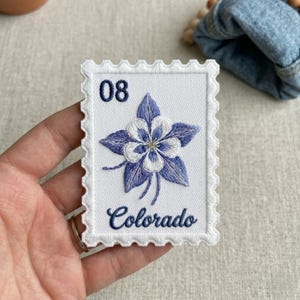 May include: Embroidered patch in the shape of a postage stamp. The patch features a blue and white flower design with the word "Colorado" embroidered below. The number "08" is embroidered in the upper left corner. The patch has a white border.