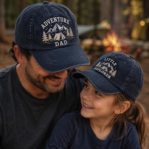 May include: Two navy blue baseball caps. One cap reads "ADVENTURE DAD" with a mountain and tent graphic. The other cap reads "LITTLE EXPLORER" with a similar graphic. Both caps are embroidered with tan thread.