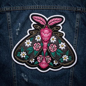 May include: Embroidered moth patch with a black background, pink body, and white and pink floral accents. The patch is outlined in white and is affixed to a denim jacket.