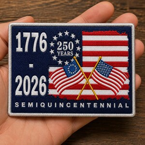 May include: A rectangular embroidered patch featuring a dark blue background with the white text 1776 and 2026, a circle of thirteen white stars surrounding the text 250 YEARS, and two crossed American flags over a red and white striped map shape.
