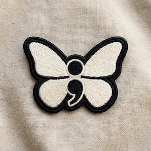 May include: Embroidered butterfly patch with a semicolon design. The butterfly has cream-colored wings with a black outline and a black semicolon in the center. The patch is on a beige fabric background.