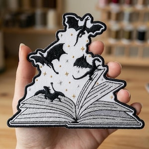 May include: Embroidered patch featuring a black and white design of a book with dragons flying out of it. The patch has a white background with black dragons and gold star accents. The book is open, with detailed stitching.