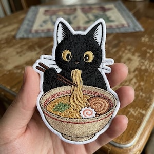 May include: Embroidered patch featuring a black cat enjoying ramen. The cat has large yellow eyes and is holding chopsticks with noodles. The ramen bowl is beige with red trim and contains noodles, vegetables, and a swirl of fish cake.