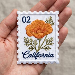 May include: Embroidered patch in the shape of a postage stamp featuring an orange poppy flower, green leaves, and the word "California" in navy blue script. The number "02" is in the upper left corner. The patch has a white border with a perforated edge.
