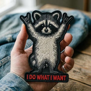 May include: Embroidered patch featuring a raccoon with its arms raised in a rock-and-roll gesture. The raccoon is black and white with a light beige chest. The patch has the text "I DO WHAT I WANT" in red letters on a black background.