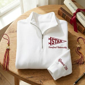 May include: A white quarter-zip sweatshirt with "STAN" and "Stanford University" embroidered in maroon. The sleeve cuff features "Grace '26" in maroon. Graduation tassels and a diploma are also visible.