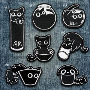 May include: A collection of seven black and white embroidered cat patches. Each patch features a different cat design, including cats in a jar, a bowl, and a tube. The patches are on a denim background.