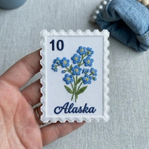 May include: Embroidered patch in the shape of a postage stamp featuring a cluster of blue forget-me-not flowers with green stems and leaves. The number "10" and the word "Alaska" are embroidered in navy blue.