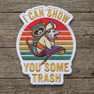 May include: Embroidered patch featuring a raccoon and opossum riding a magic carpet against a sunset backdrop. The patch reads "I CAN SHOW YOU SOME TRASH" in gold lettering, with a white border.