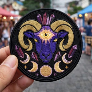 May include: Embroidered patch featuring a purple ram's head with golden horns, an all-seeing eye, and crystal accents. The design includes crescent moons, stars, and a large golden circle on a black background. The patch is round.