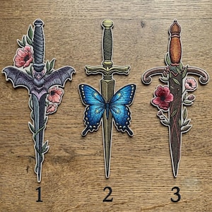 May include: Three decorative dagger patches with floral and insect designs. One features a bat, another a blue butterfly, and the third has flowers. Each patch has a number at the bottom. The daggers have ornate handles and blades.