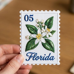 May include: Embroidered patch in the shape of a postage stamp. The patch features a floral design with white flowers, green leaves, and the word "Florida" in blue script. The number "05" is in the upper left corner.