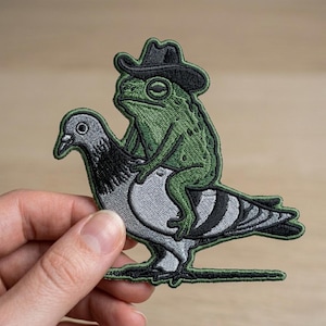 May include: Embroidered patch featuring a green frog wearing a black cowboy hat, riding a gray pigeon. The patch has a black border and is held against a light brown background. The frog is the focal point, with detailed stitching.