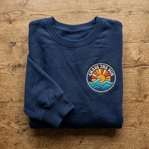 May include: Navy blue sweatshirt with a round embroidered patch featuring a sun, waves, and the text "CHASE THE SUN". The sweatshirt is folded on a wooden surface.