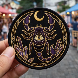May include: Round black patch featuring a purple scorpion with a crescent moon, eye, and crystal design. The patch has gold and purple accents and is likely an iron-on or sew-on patch for clothing or accessories.