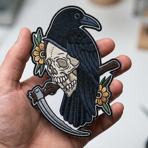 May include: Embroidered patch featuring a black crow perched atop a skull with a scythe. The design includes yellow flowers and green leaves. The patch is detailed with shades of black, white, brown, and yellow.