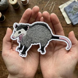 May include: Embroidered patch featuring a gray opossum with a pink mouth and tail, black paws and ears, and a white border. The opossum is in a walking pose with its mouth open. The patch is held in open hands.