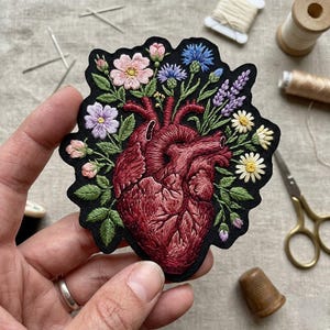 May include: Embroidered patch featuring a detailed anatomical heart design, bursting with colorful flowers and green leaves. The heart is a deep red, set against a black background. The patch is surrounded by sewing supplies, including needles, thread, and scissors.