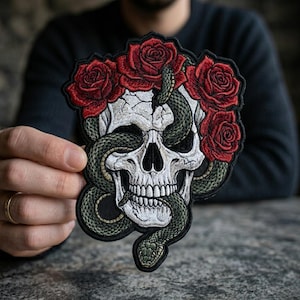 May include: Embroidered patch featuring a white skull with a snake entwined around it. Red roses form a crown above the skull. The patch has a black border and is held up by a person's hand.