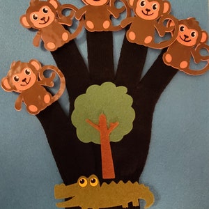 May include: A black glove with five brown monkey finger puppets. A green felt tree and alligator are also attached. The background is light blue. This is a children's toy.