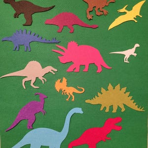 May include: Assortment of colorful felt dinosaur cutouts in various shapes and sizes, including a T-Rex, Triceratops, and Stegosaurus. The dinosaurs are in shades of brown, pink, blue, purple, yellow, and orange, set against a green background.
