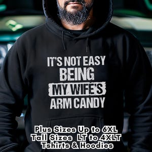 Funny Gift for Husband - this image shows a bearded man wearing a black pullover Hoodie sweatshirt with text saying "It's not easy being my wife's arm candy" - image includes a text overlay saying that we carry Shirts and Hoodies in Big sizes 3XL, 4XL, 5XL, and 6XL and Tall sizes LT, XLT, 2XLT, 3XLT, and 4XLT