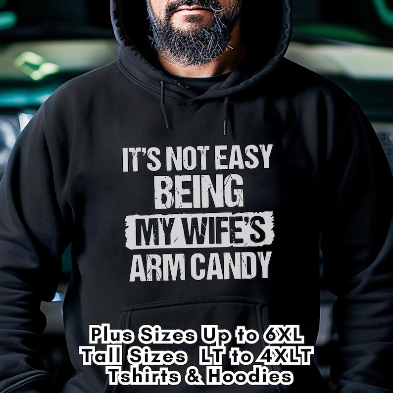 Funny Gift for Husband - this image shows a bearded man wearing a black pullover Hoodie sweatshirt with text saying "It's not easy being my wife's arm candy" - image includes a text overlay saying that we carry Shirts and Hoodies in Big sizes 3XL, 4XL, 5XL, and 6XL and Tall sizes LT, XLT, 2XLT, 3XLT, and 4XLT