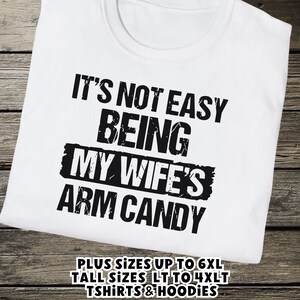Funny Gift for Husband - a tshirt with text saying "It's not easy being my wife's arm candy" on a White shirt - image includes a text overlay saying that we carry Shirts and Hoodies in Big sizes 3XL, 4XL, 5XL, and 6XL and Tall sizes LT, XLT, 2XLT, 3XLT, and 4XLT