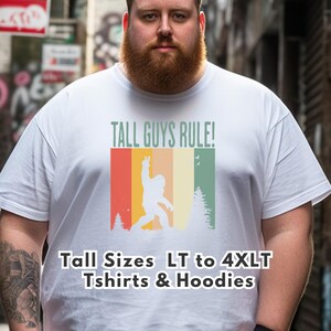 Puede incluir: Camiseta blanca con el texto "TALL GUYS RULE!" y un gr&aacute;fico retro de un Bigfoot haciendo el signo de la paz. El gr&aacute;fico tiene rayas rojas, naranjas, amarillas y verdes. La camiseta tambi&eacute;n tiene el texto "Tall Sizes LT to 4XLT Tshirts & Hoodies".