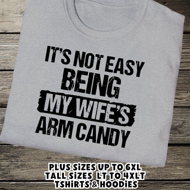 Funny Gift for Husband - a tshirt with text saying "It's not easy being my wife's arm candy" on a Sport Grey shirt - image includes a text overlay saying that we carry Shirts and Hoodies in Big sizes 3XL, 4XL, 5XL, and 6XL and Tall sizes LT, XLT, 2XLT, 3XLT, and 4XLT