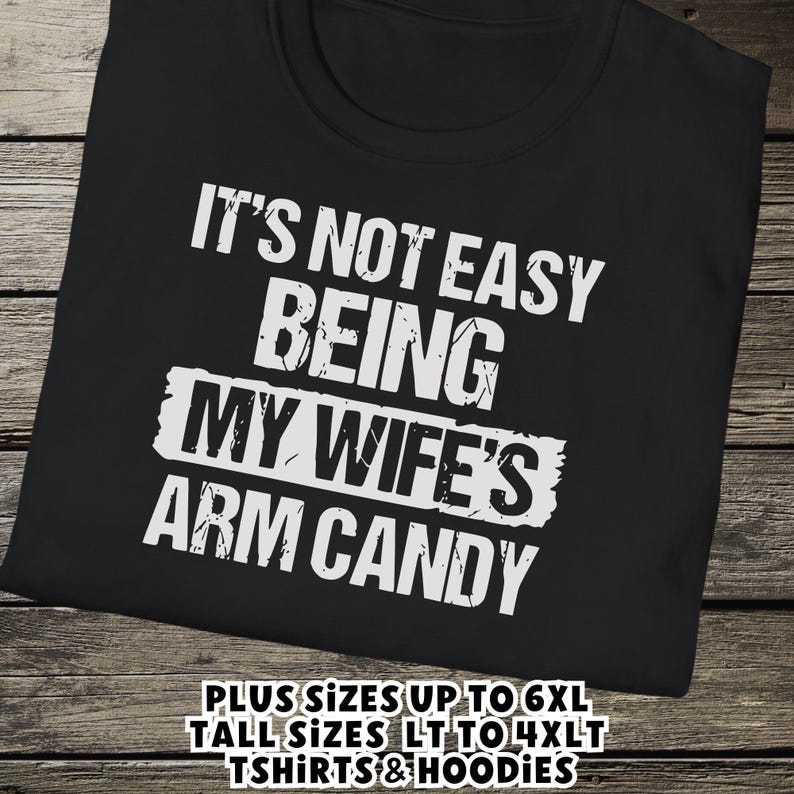 Funny Gift for Husband - a tshirt with text saying "It's not easy being my wife's arm candy" on a Black shirt - image includes a text overlay saying that we carry Shirts and Hoodies in Big sizes 3XL, 4XL, 5XL, and 6XL and Tall sizes LT, XLT, 2XLT, 3XLT, and 4XLT