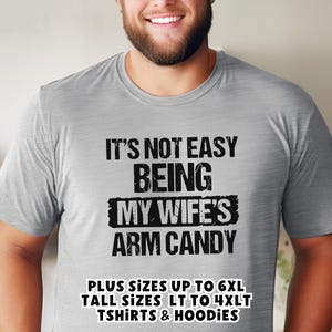 Funny Gift for Husband - a young man wearing a sport grey Tshirt with text saying "It's not easy being my wife's arm candy" - image includes a text overlay saying that we carry TShirts and Hoodies in Big sizes 3XL, 4XL, 5XL, and 6XL and Tall sizes LT, XLT, 2XLT, 3XLT, and 4XLT