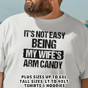 May include: White t-shirt with the text "IT'S NOT EASY BEING MY WIFE'S ARM CANDY" in black. The shirt also states "PLUS SIZES UP TO 6XL, TALL SIZES LT TO 4XLT, TSHIRTS & HOODIES".