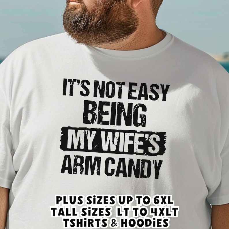Funny Gift for Husband - a bearded plus size big guy wearing a white Tshirt with black vintage text saying "It's not easy being my wife's arm candy" - image includes a text overlay saying that we carry TShirts and Hoodies in Big sizes 3XL, 4XL, 5XL, and 6XL and Tall sizes LT, XLT, 2XLT, 3XLT, and 4XLT