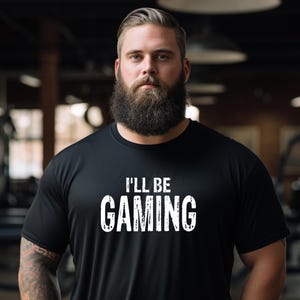 May include: Black t-shirt with the white text "I'LL BE GAMING". The shirt is made of a soft material. The person in the image has a full beard and tattoos on their arm.