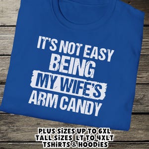 Funny Gift for Husband - a tshirt with text saying "It's not easy being my wife's arm candy" on a Royal Blue shirt - image includes a text overlay saying that we carry Shirts and Hoodies in Big sizes 3XL, 4XL, 5XL, and 6XL and Tall sizes LT, XLT, 2XLT, 3XLT, and 4XLT