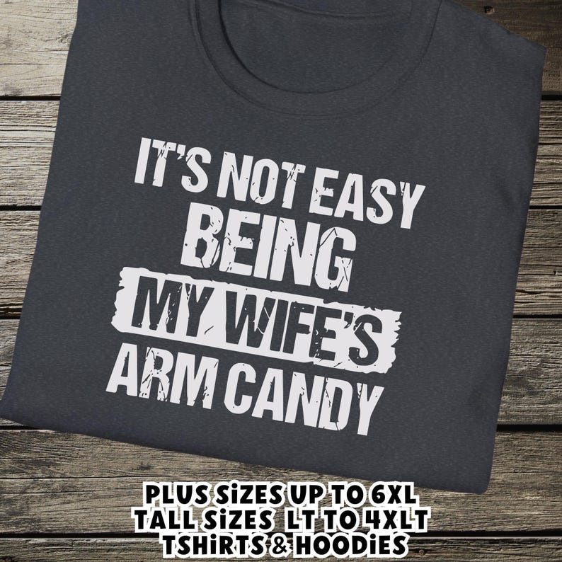 Funny Gift for Husband - a tshirt with text saying "It's not easy being my wife's arm candy" on a Dark Grey Heather shirt - image includes a text overlay saying that we carry Shirts and Hoodies in Big sizes 3XL, 4XL, 5XL, and 6XL and Tall sizes LT, XLT, 2XLT, 3XLT, and 4XLT