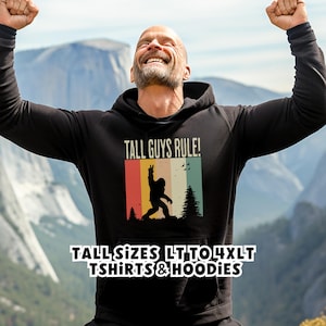 Puede incluir: Sudadera con capucha negra con el texto "TALL GUYS RULE!" y un gr&aacute;fico de Bigfoot en un dise&ntilde;o retro de bloques de color. La sudadera tambi&eacute;n tiene el texto "TALL SIZES LT TO 4XLT TSHIRTS & HOODIES".