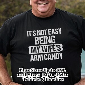 Funny Gift for Husband - a plus size older man wearing a black Tshirt with text saying "It's not easy being my wife's arm candy" - image includes a text overlay saying that we carry TShirts and Hoodies in Big sizes 3XL, 4XL, 5XL, and 6XL and Tall sizes LT, XLT, 2XLT, 3XLT, and 4XLT