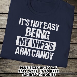Funny Gift for Husband - a tshirt with text saying "It's not easy being my wife's arm candy" on a Navy Blue shirt - image includes a text overlay saying that we carry Shirts and Hoodies in Big sizes 3XL, 4XL, 5XL, and 6XL and Tall sizes LT, XLT, 2XLT, 3XLT, and 4XLT