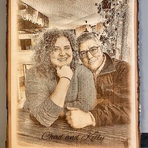 May include: A rectangular live-edge wood plaque featuring a laser-engraved portrait of two people sitting at a table. The engraving shows a person with curly hair and a person wearing glasses. The text Chad and Kelly is engraved at the bottom.