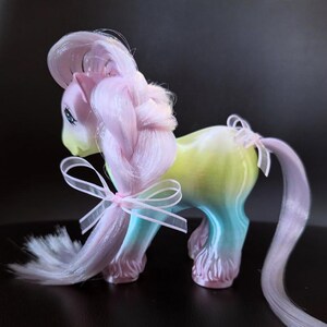 May include: A pastel-colored toy pony with a braided mane and tail, adorned with delicate pink bows. The pony's body transitions from yellow to blue, with pink hooves and a matching pink mane. The toy has blue eyes and a light yellow face.